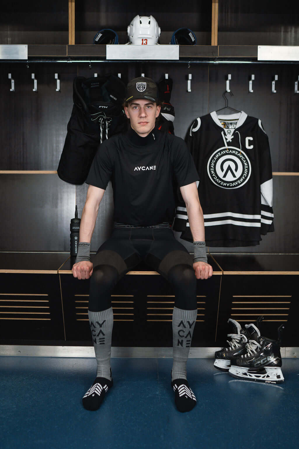 Hockey player wearing AVCAne gear, seated in a locker room with hockey equipment and jerseys.