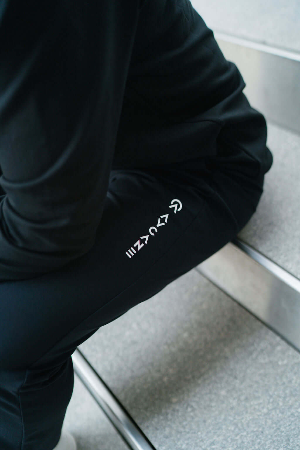 Close-up of a person wearing Auric Pants on stairs, showcasing ultra-stretchy fabric and stylish logo design.