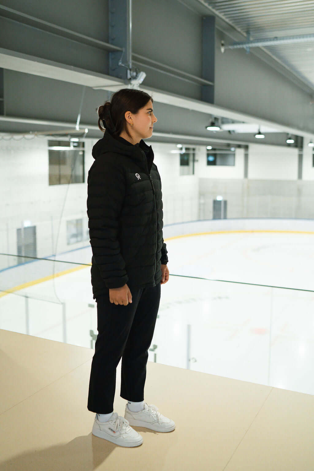 Woman wearing a black T-Lite Jacket, standing near an ice rink, showcasing warmth and stylish design.