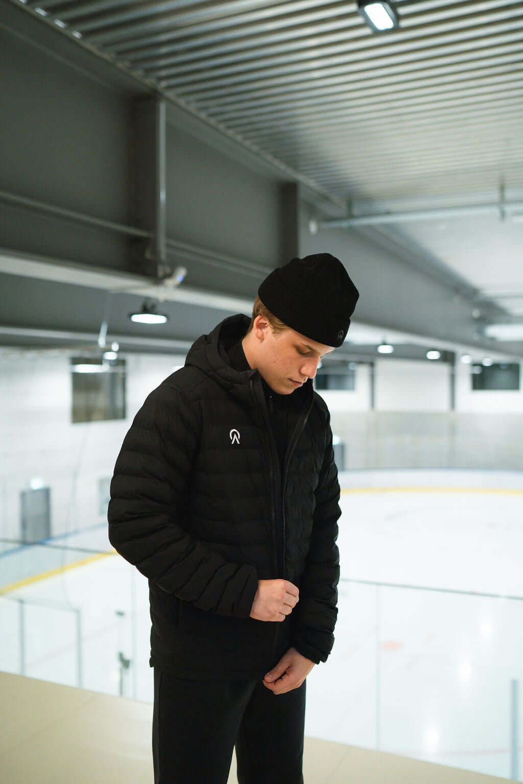 Young man wearing a black T-Lite Jacket and hat indoors near an ice rink, showcasing warmth and style.