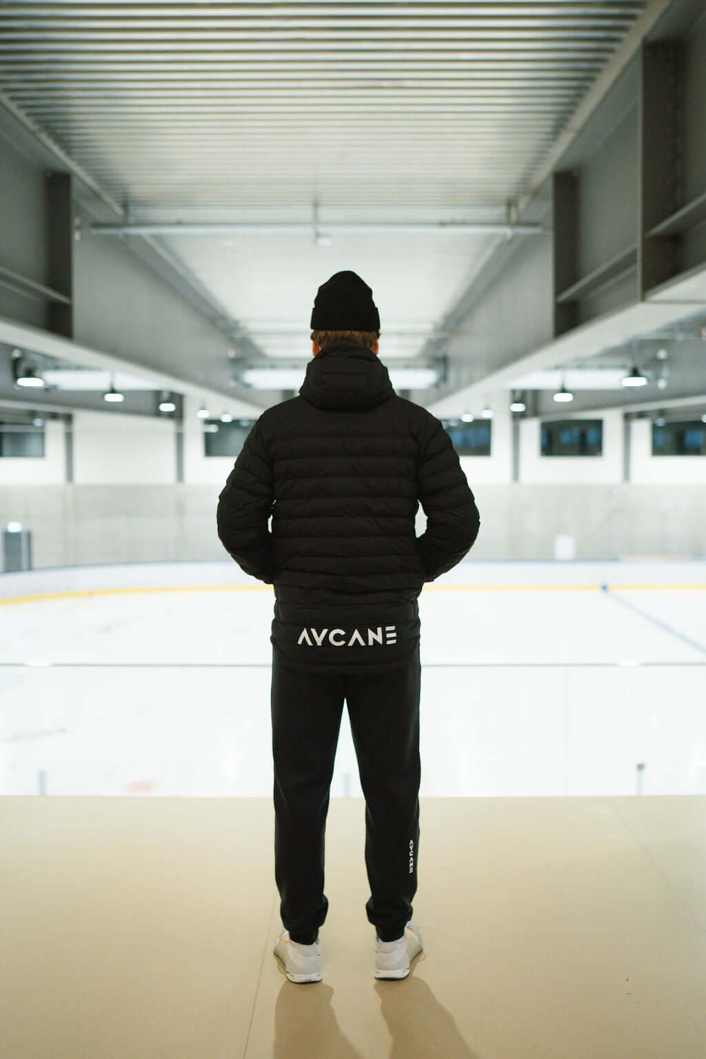 Person in a black T-Lite Jacket and pants, standing on a balcony overlooking an ice rink.