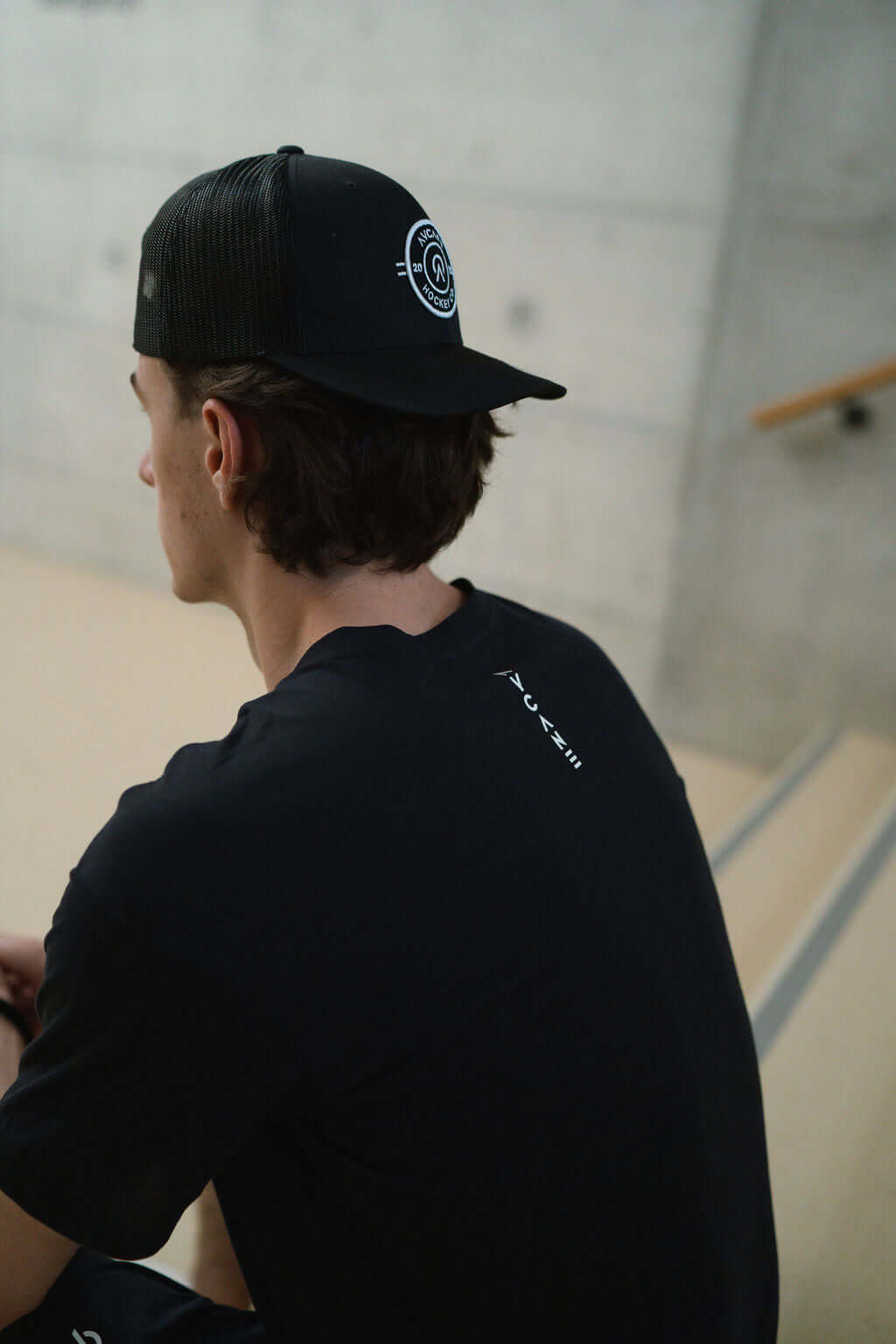 Young man wearing a black Route Short Sleeve T-Shirt and a cap, seated on stairs in a modern space.