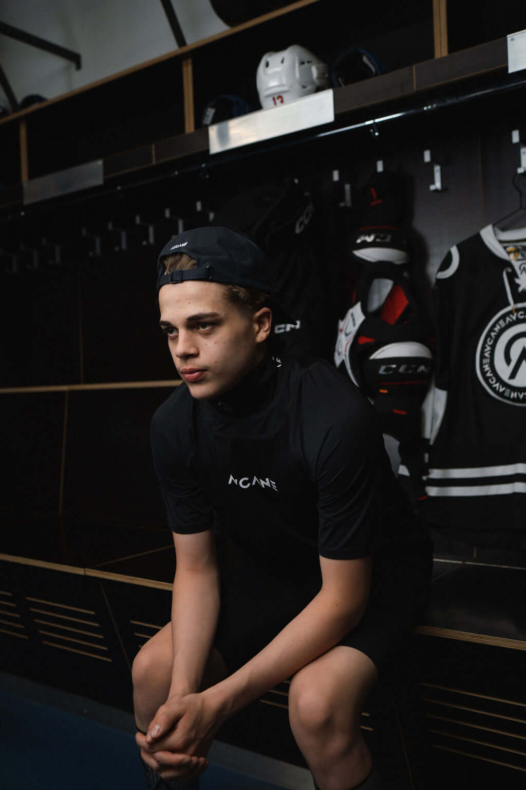 Young athlete in a locker room wearing a black short sleeve neck guard shirt, looking thoughtful, surrounded by hockey gear.