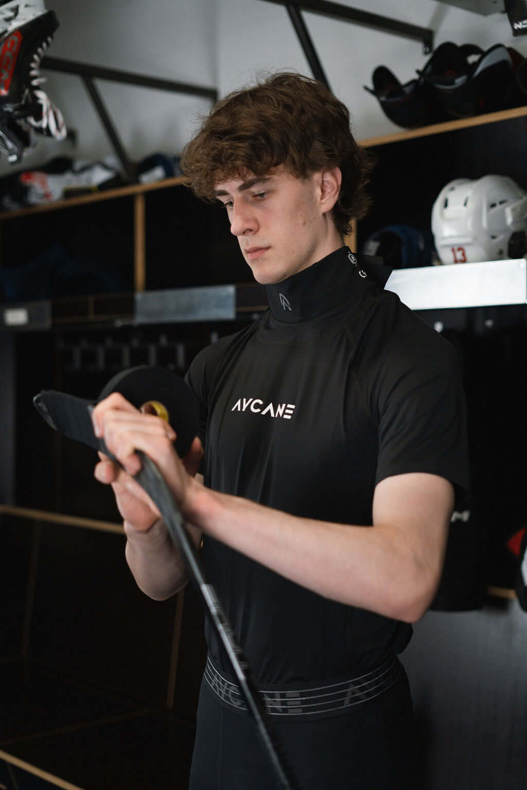 Athlete wearing Blade X short sleeve base layer, preparing hockey stick in locker room.