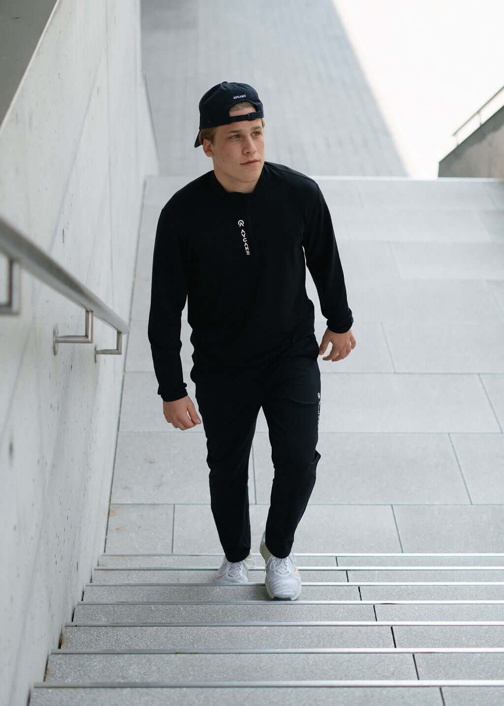 Man wearing a black Maurize midlayer and pants, walking up urban stairs, showcasing activewear style.