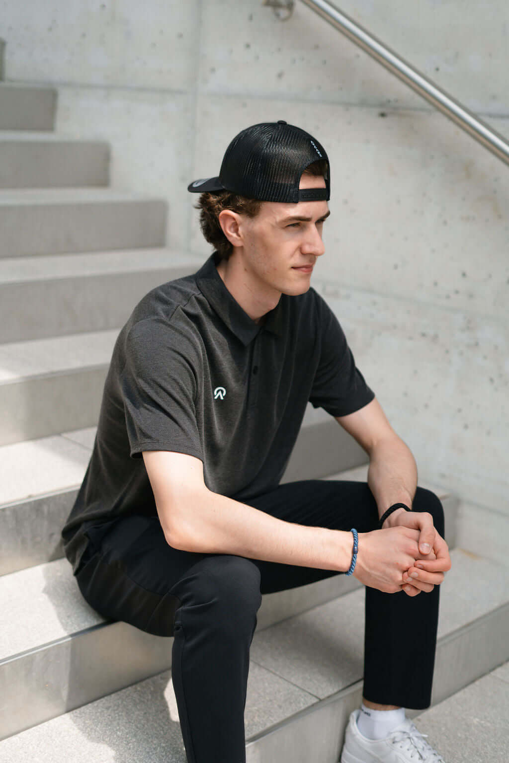 Young man in a black Netter Polo shirt seated on stairs, wearing a cap and casual pants.