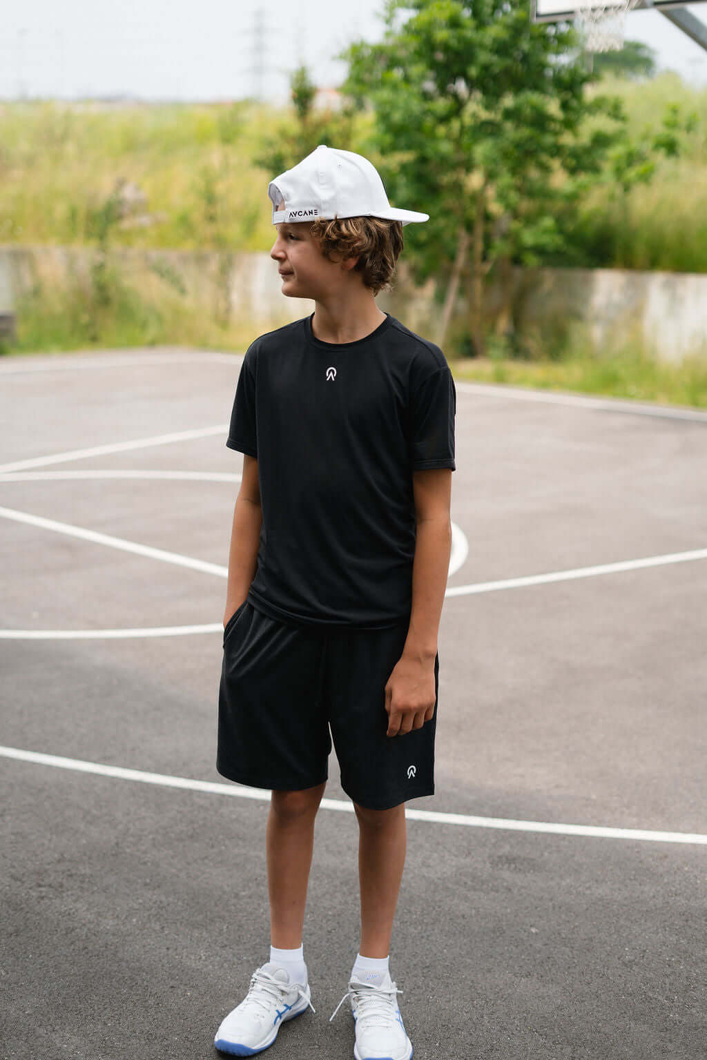 Young boy in a black Route Short Sleeve T-Shirt and shorts on a basketball court, wearing a cap.