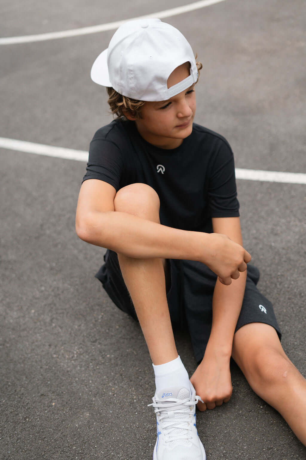 Young boy wearing a black Route T-Shirt and cap, sitting on a basketball court, showcasing comfort and style.