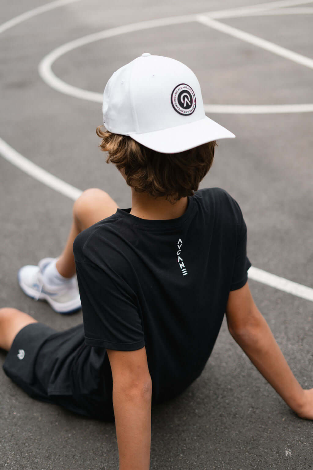 Young boy wearing a black Route Short Sleeve T-shirt and white cap, sitting on a basketball court.