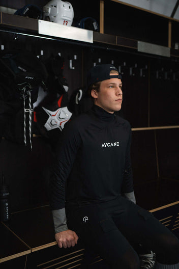Young athlete wearing a black long sleeve base layer shirt, seated in a locker room, preparing for hockey.