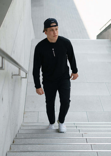 Man wearing a black Maurize midlayer and pants, walking up urban stairs, showcasing activewear style.