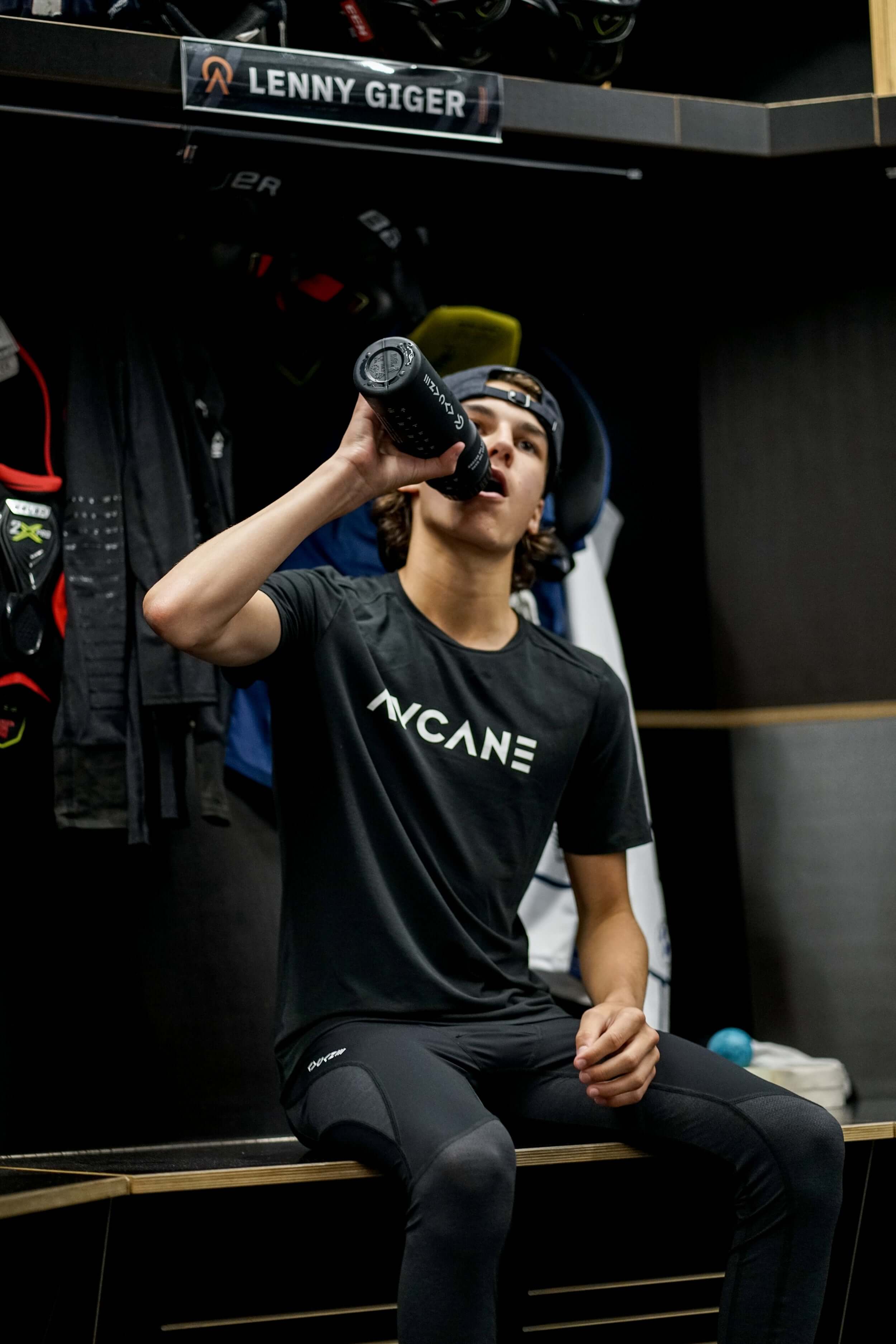 Youth hockey player in locker room drinking from a AYCANE water bottle wearing a short sleeve training t-shirt with a big white AYCANE logo on chest